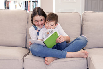Smiling tween girl reading book to her little brother sitting in her arms with soft furry rabbit toy on sofa at home. Siblings care and love. Sister and brother quality time.