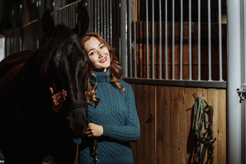 Young woman in sweater readying black horse for a ride while standing inside a stable on a farm