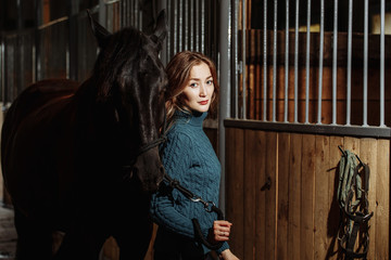 Young woman in sweater readying black horse for a ride while standing inside a stable on a farm