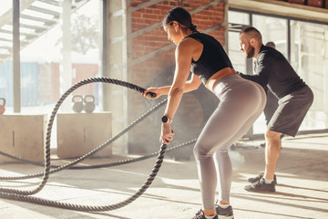 Fototapeta na wymiar Caucasian fit attractive couple in sports clothing concentrated on the exercise with battle rope requiring increased effort and strength, standing at well lit fitness centre with loft brick interior