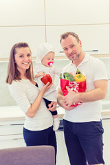 Happy parents standing in the kitchen and prepares for cooking with their baby girl chef. Father bring vegetables in paper bag.