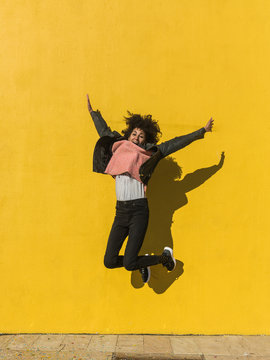 Black woman with afro hair jumping for joy in the street with a yellow wall in the background