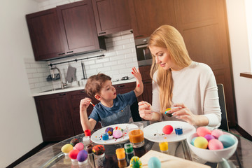 Cute little male kid shows his young mother his small palms,covered with paint after decorating Easter eggs. Mother and son preparing to Easter at kitchen, painting.