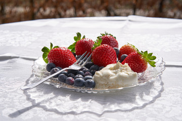Light juicy fruit dessert ideal for diet consist from strawberries, bluberries and sour cream, served on the small glass plate with fork on the table of garden restaurant or cafe in summer sunny day.