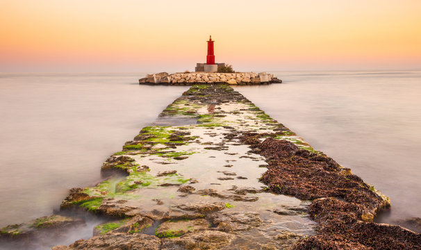Damaged stone path going in calm sea water towards small islet with beacon during wonderful sunrise