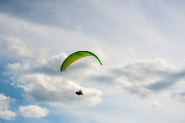 Yellow and green paraglider is flying in the blue sky against the background of clouds. Paragliding in the sky on a sunny day.