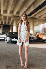 Young positive sexy caucasian girl driver in elegant stylish dress on parking outdoors smiling 