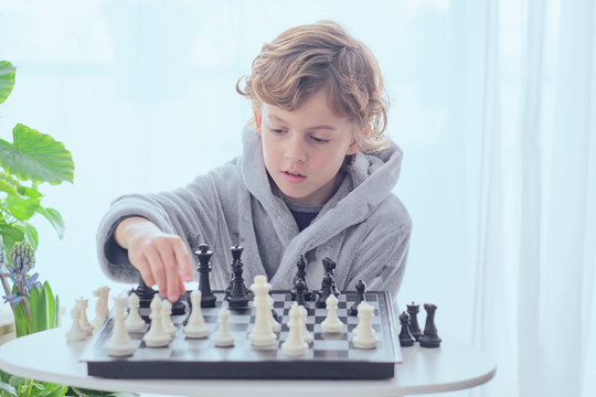 Smart Child Holding White Figure On Chess Board At Table Near White Curtains