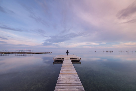 Fototapeta Back view of unrecognizable person standing on long lumber pier near calm sea water on cloudy evening in Spain