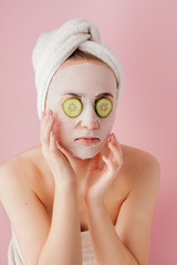 Beautiful young woman is applying a cosmetic tissue mask on a face with cucumber on a pink background