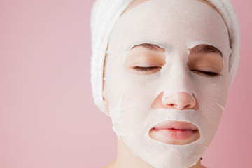 Beautiful young woman is applying a cosmetic tissue mask on a face on a pink background. Healthcare and beauty treatment and technology concept