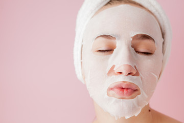Beautiful young woman is applying a cosmetic tissue mask on a face on a pink background. Healthcare and beauty treatment and technology concept