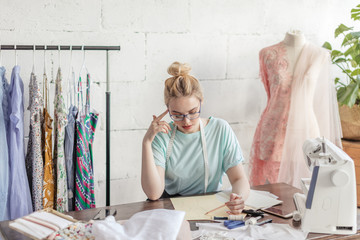 Pensive beautiful blonde dressmaker in fashion atelier in stylish eyeglasses and mint casual...