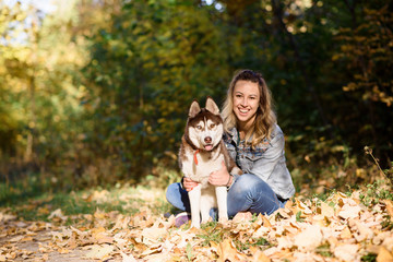 Naklejka na ściany i meble Smiling young brunette girl sitting with her husky dog in green park outdoors. Cute and friendly couple enjoying the nature. Pretty female and fluffy doggy