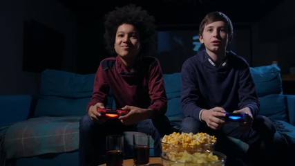 Diverse casually dressed teenagers with game controllers fighting in shooter game on home console sitting side by side on sofa. Winner always differs from loser, emotions written on face