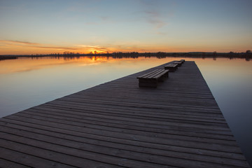 A wide wooden jetty with benches, sunset and calm lake