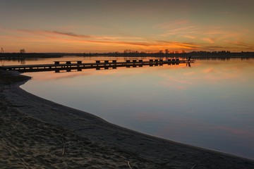 Sandy shore of a calm lake, jetty and clouds after sunset