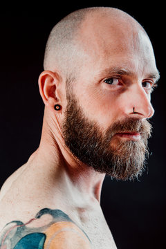 Side view of bald thoughtful hipster with earring and piercing looking away on black background