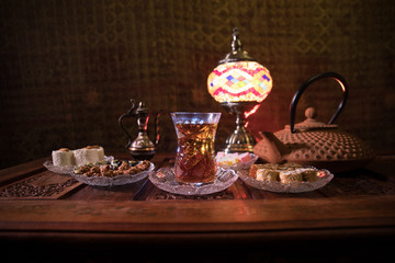 Arabian tea in glass with eastern snacks on vintage wooden surface. Eastern tea concept. Low light lounge interior with carpet. Empty space.