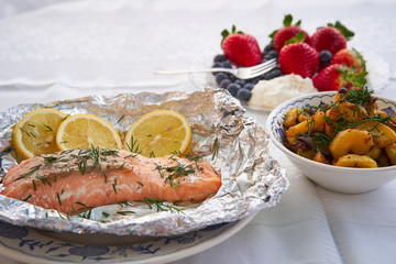 Salmon fillet baked in aluminium foil with slices of organic lemon and chopped fresh dill served with bowl of roasted potatoes and onions and small plate with fresh blueberries, strawberries and cream
