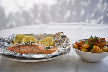 Salmon fillet or steak baked in aluminium foil with slices of organic lemon and chopped fresh dill served with bowl of roasted potatoes with onion, fresh, healty, low fat food, nutrition balanced diet