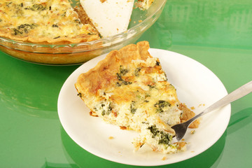 Slice of bacon and spinach quiche on white plate with pie plate in BG