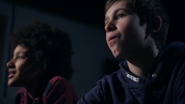 Close up teenager eating popcorn looking at TV screen. Chewing caucasian boy watching comic film with mixed race friend blurry on background and laughing viewing especially comical scenes of movie