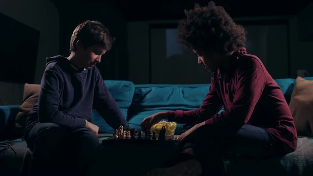 Smart teenagers playing chess game on boad sitting on sofa in dark living room at night. Each of diverse friends makes move by chess piece, carefully thought out, no one wants to be a loser