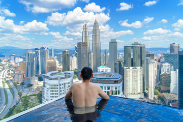 Back of tourist in a swimming pool on rooftop with Kuala Lumpur downtown view and blue sky. Malaysia travel trip in vacation and holidays concept in Asia. Skyscraper and high-rise buildings at noon. - obrazy, fototapety, plakaty