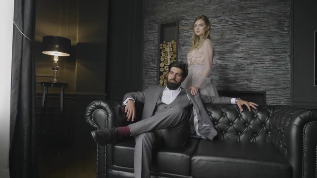 slow motion severe bearded man in grey suit sits lolling on leather sofa blond lady stands behind by decorated wall