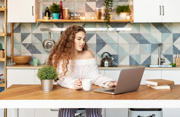 Portrait of young blogger girl using modern laptop and wearing comfy knitted white sweater. Sweet curly lady holding cup of tea. Technology and nice interior concept