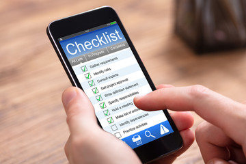 Person Filling Checklist Form On Cellphone At Wooden Desk