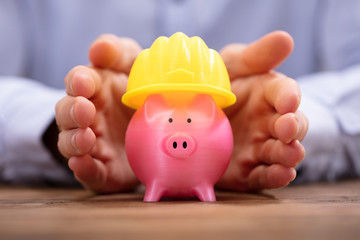 Person Protecting Piggy Bank With Yellow Hard Hat