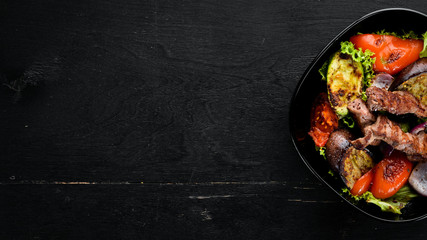 Baked beef and grilled vegetables in a black plate. On a wooden background. Top view. Free space for your text.