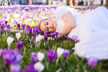 Obraz na płótnie Canvas Beautiful young blonde woman with blue eyes wearing in white dress lying on the carpet among the spring flowers crocuses. Spring sunny day. A field of purple flowers. Springtime saffron field