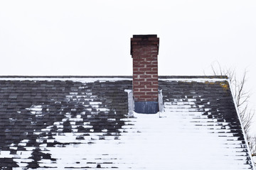Brown brick chimney on grey asphalt shingles roof partly covered with snow in winter against sky with copy space for text.
