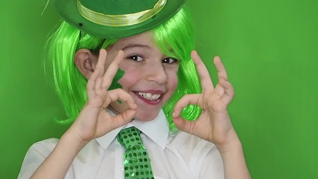 Child Celebrating St. Patrick's Day Showing his Make-up. A small, boy in green wig with leaf of clover and Irish flag on his cheeks showing a gesture okay. slow motion. green background