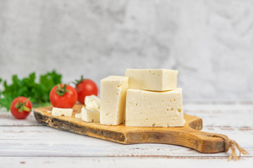 Large pieces of feta cheese on old wooden cutting board and cherry tomatoes on light background. Selective focus,
