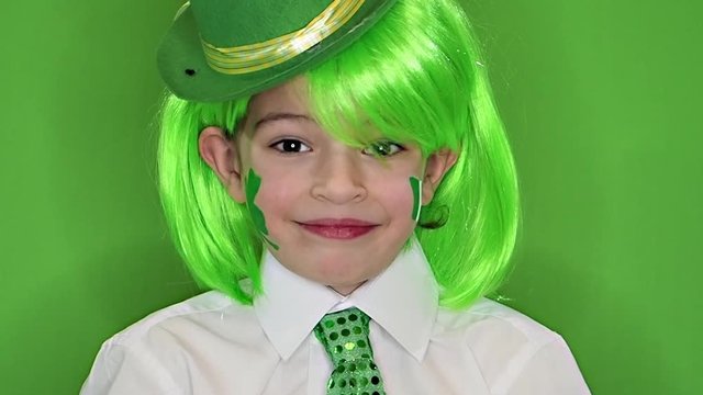 Child Celebrating St. Patrick's Day Showing his Make-up. A small, boy in green carnival wig with drawing in the form of a leaf of clover on his cheek. slow motion. green background