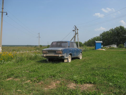 Coche antiguo en un parado ruso. Fondo hierba verde y arboles y azules manches de coche, casita y cielo