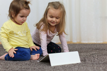 Two beautiful little sisters sit at a table and play on a tablet
