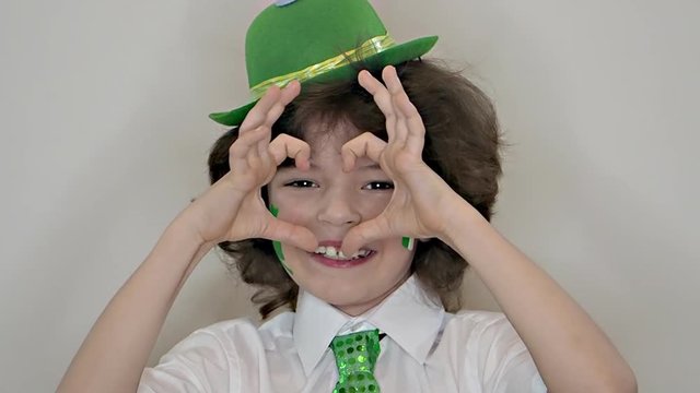 Child Celebrating St. Patrick's Day Showing his Make-up. A small, curvy boy leaf of clover and Irish flag on his cheeks showing a gesture of love. slow motion. light background
