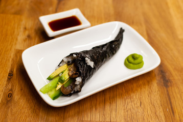 Homemade vegan Temaki-Sushi filled with avocado on a small plate on a wooden table with soy sauce and wasabi