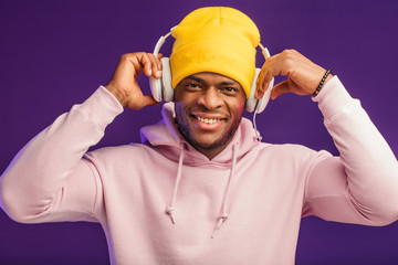 Joyful happy hipster man in white hoodie and yellow hat listening music with headphones, isolated at purple studio background. Hip hop style, positive emotions, face expression, dancing concept