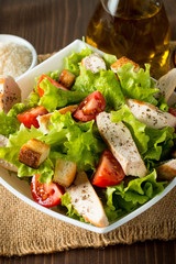 Fresh salad made of tomato, ruccola, chicken breast, eggs, arugula, crackers and spices. Caesar salad in a white, transparent bowl on wooden background