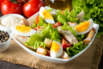 Fresh salad made of tomato, ruccola, chicken breast, eggs, arugula, crackers and spices. Caesar salad in a white, transparent bowl on wooden background