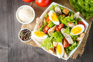 Fresh salad made of tomato, ruccola, chicken breast, eggs, arugula, crackers and spices. Caesar salad in a white, transparent bowl on wooden background