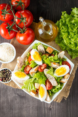 Fresh salad made of tomato, ruccola, chicken breast, eggs, arugula, crackers and spices. Caesar salad in a white, transparent bowl on wooden background
