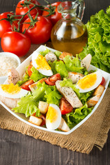 Fresh salad made of tomato, ruccola, chicken breast, eggs, arugula, crackers and spices. Caesar salad in a white, transparent bowl on wooden background
