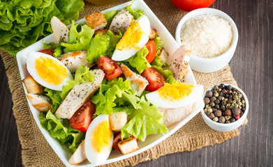 Fresh salad made of tomato, ruccola, chicken breast, eggs, arugula, crackers and spices. Caesar salad in a white, transparent bowl on wooden background